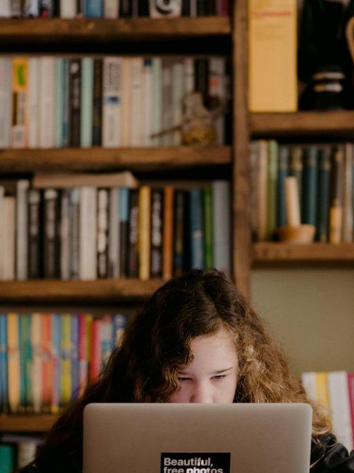 Ein Mädchen sitzt vor einer Bücherwand vor seinem Laptop.