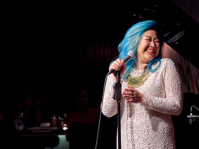 Die Sängerin Bihzhu, blau gefärbtes Haar, weißes Spitzenkleid, am Mikrofon auf der Bühne - sie schaut zur Seite und lacht