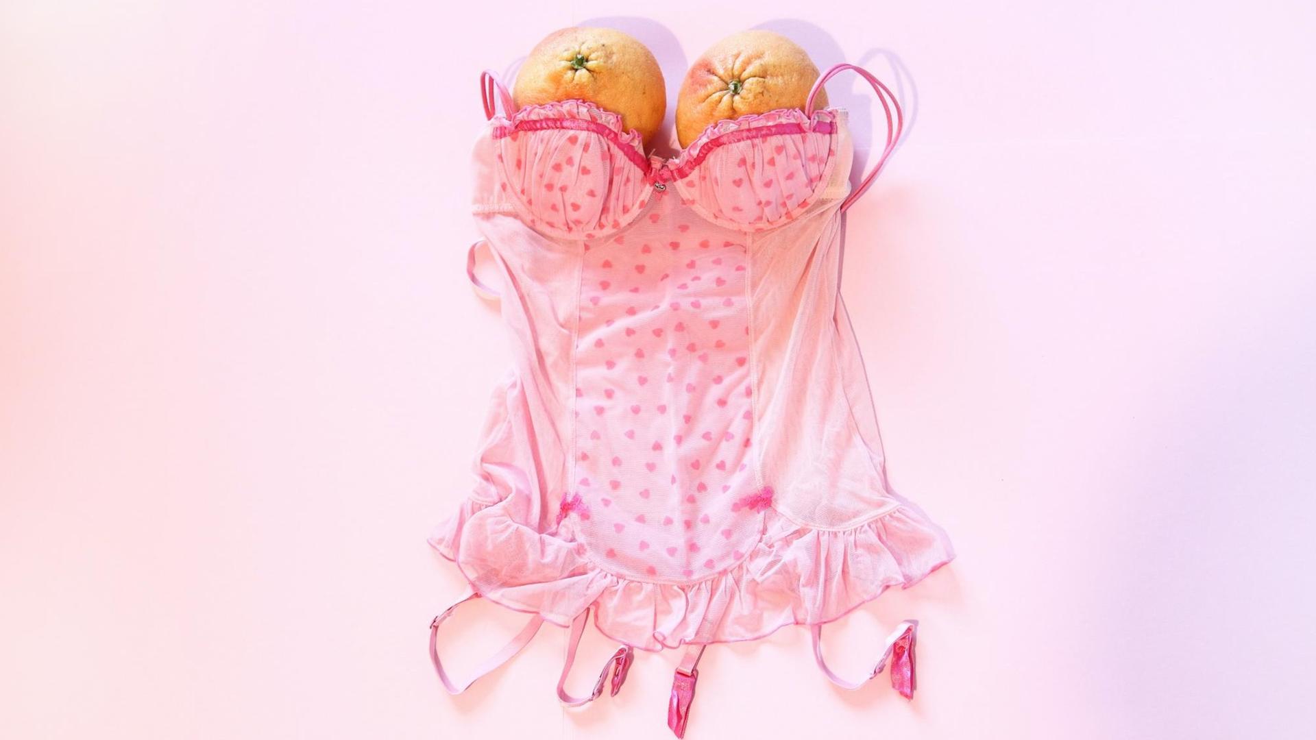 Eine rosa Korsage liegt auf rosa Hintergrund – dort wo beim Tragen die Brüste wären, sind zwei Orangen platziert. Eine rosa Korsage liegt auf rosa Hintergrund – dort wo beim Tragen die Brüste wären, sind zwei Orangen platziert.
