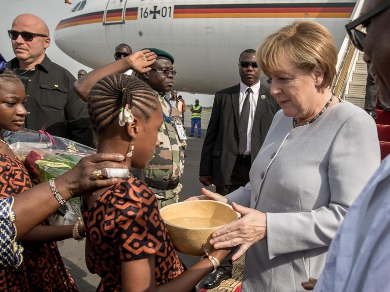 Bundeskanzlerin Angela Merkel (CDU) wird am 09.10.2016 in Bamako in Mali neben Staatspräsident Ibrahim Boubacar Keita (r) am Flughafen von Blumenmädchen begrüßt, die der Kanzlerin zunächst eine traditionelle Wasserkalebasse reichen.