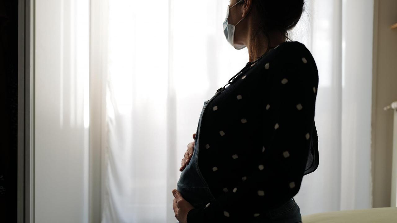 Eine schwangere Frau sitzt mit Gesichtsmaske auf einem Bett und blickt melancholisch aus dem Fenster. Eine schwangere Frau sitzt mit Gesichtsmaske auf einem Bett und blickt melancholisch aus dem Fenster.