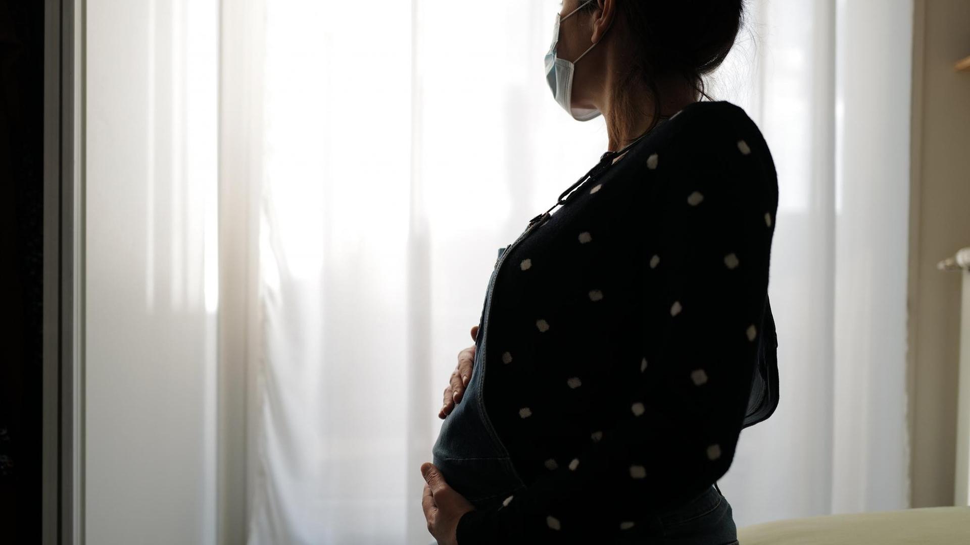 Eine schwangere Frau sitzt mit Gesichtsmaske auf einem Bett und blickt melancholisch aus dem Fenster. Eine schwangere Frau sitzt mit Gesichtsmaske auf einem Bett und blickt melancholisch aus dem Fenster.