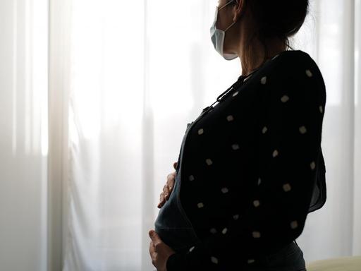 Eine schwangere Frau sitzt mit Gesichtsmaske auf einem Bett und blickt melancholisch aus dem Fenster.