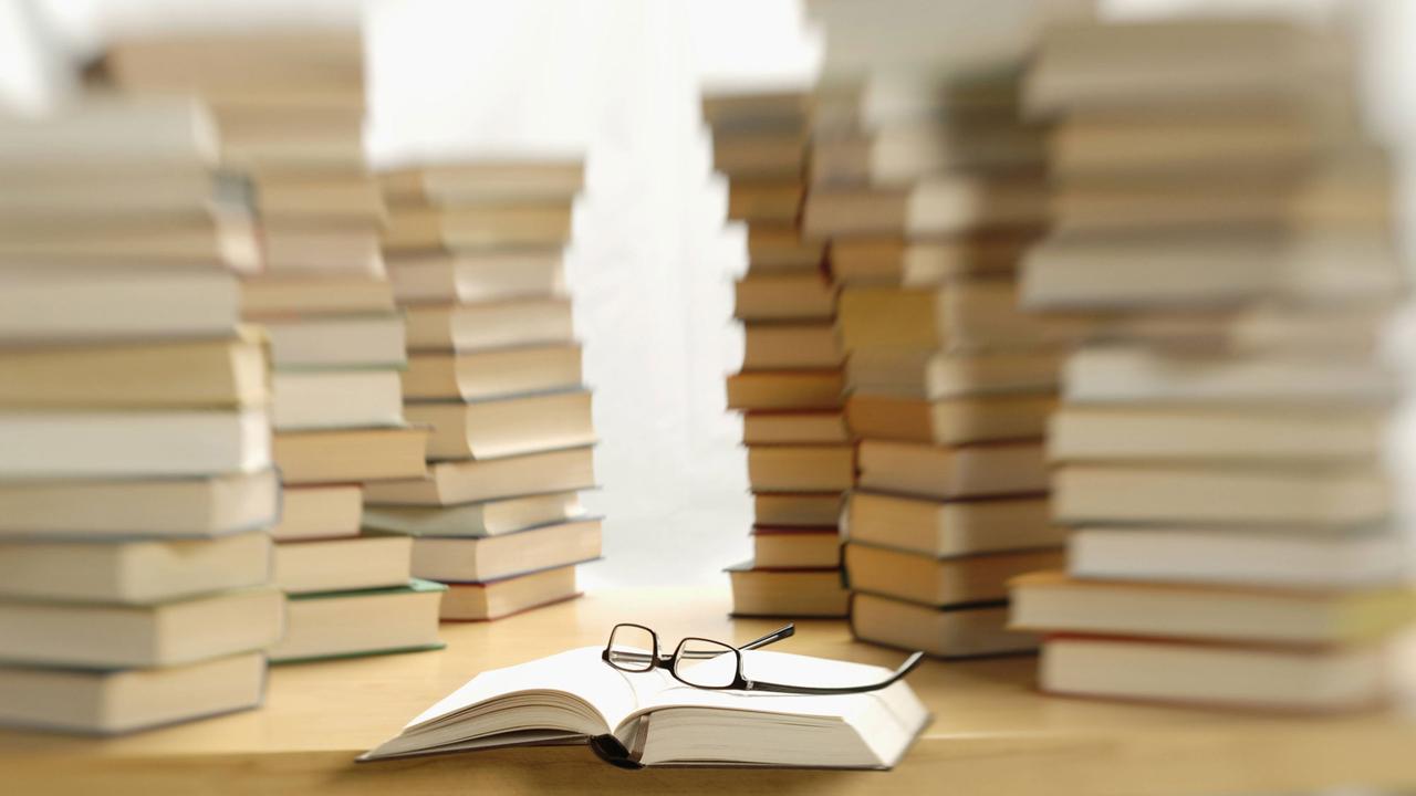 Mehrere Bücherstapel auf einem Tisch, davor ein aufgeschlagenes Buch, auf dem eine Brille liegt Mehrere Bücherstapel auf einem Tisch, davor ein aufgeschlagenes Buch, auf dem eine Brille liegt