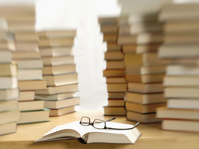 Mehrere Bücherstapel auf einem Tisch, davor ein aufgeschlagenes Buch, auf dem eine Brille liegt