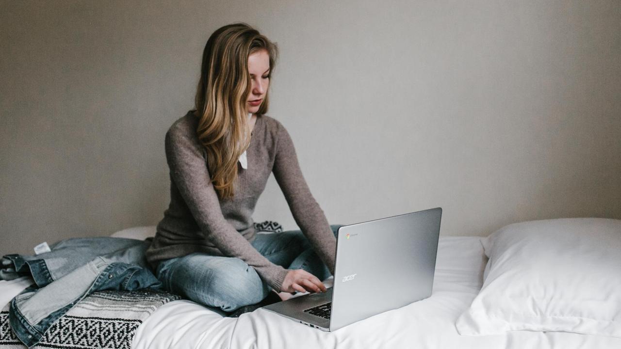 Eine junge Frau sitzt mit ihrem Laptop auf dem Bett.