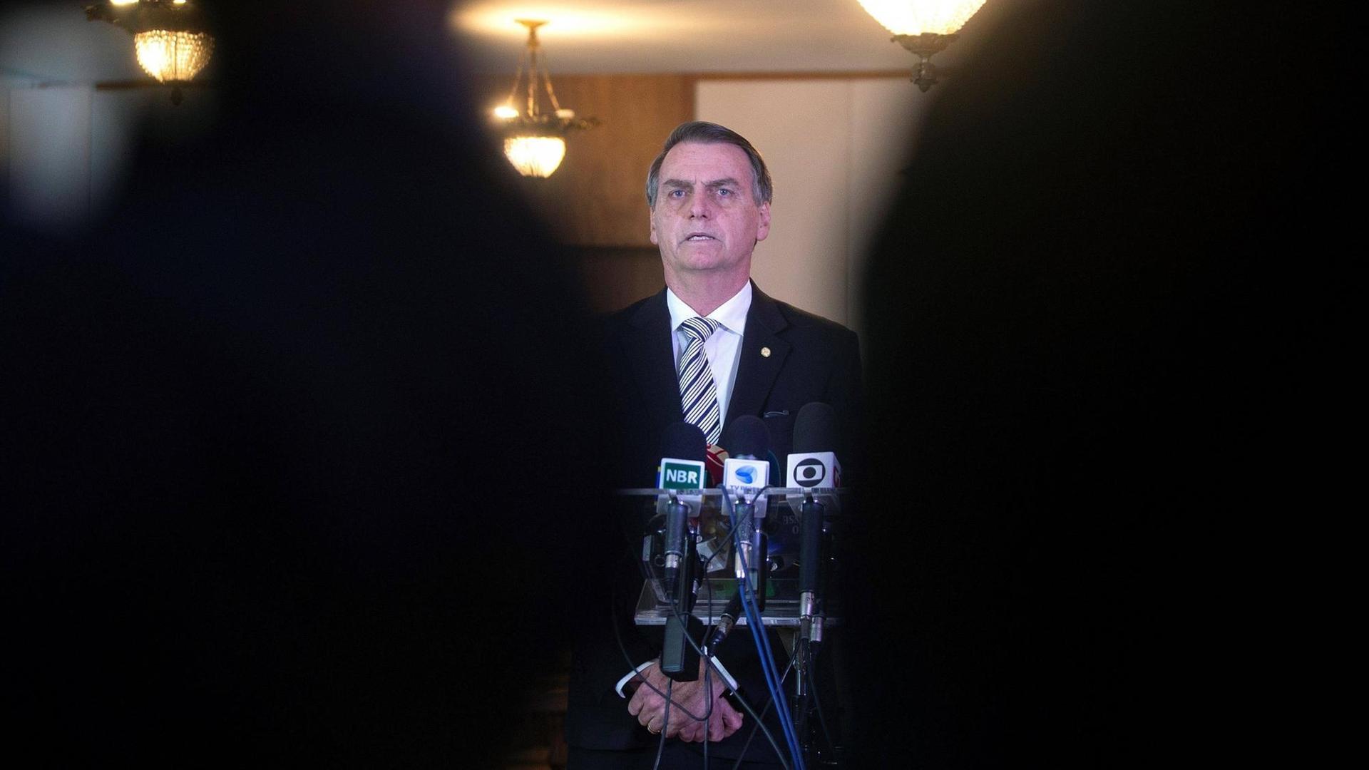 Durch eine schmale Lücke zwischen zwei Personen im Vordergrund ist Brasiliens Präsident Jair Bolsonaro zu sehen. Vor ihm stehen mehrere Mikrofone. Durch eine schmale Lücke zwischen zwei Personen im Vordergrund ist Brasiliens Präsident Jair Bolsonaro zu sehen. Vor ihm stehen mehrere Mikrofone.