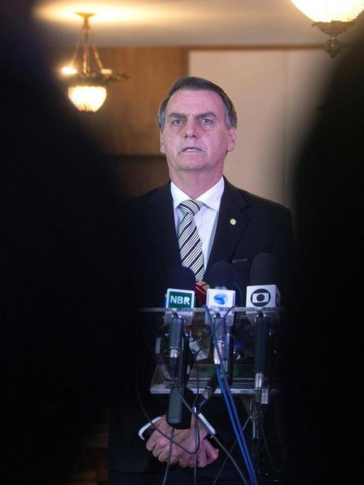 Durch eine schmale Lücke zwischen zwei Personen im Vordergrund ist Brasiliens Präsident Jair Bolsonaro zu sehen. Vor ihm stehen mehrere Mikrofone.