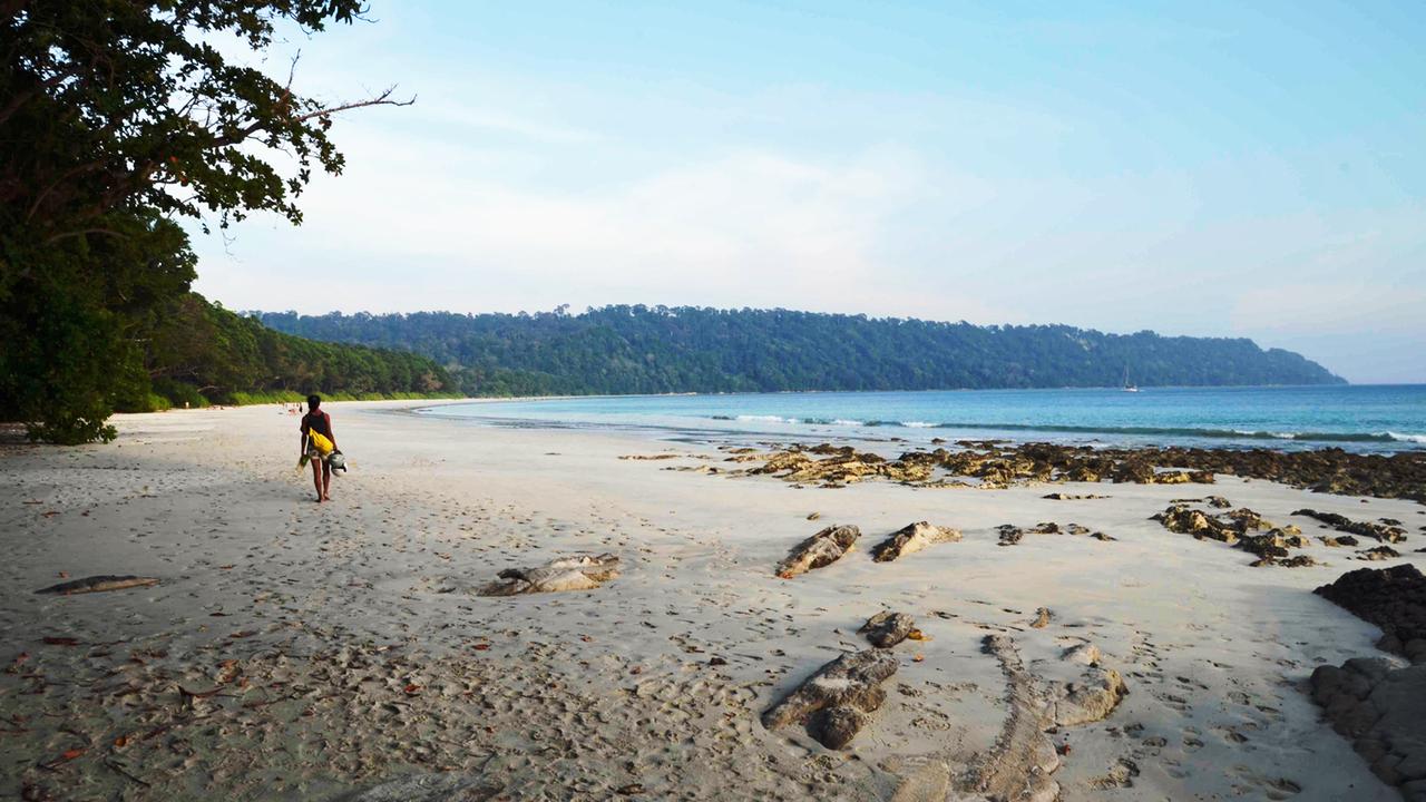Ein Tourist mit Gepäck. läuft einen breiten Sandstrand am Meer entlang. Im Hintergrund die Wälder der Andamanen.