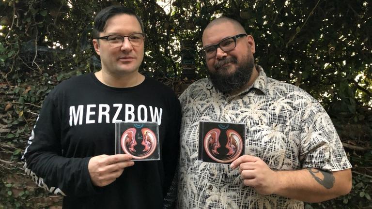 Auf dem Bild sind die Macher des Merzcasts Mike Connelly und Greh Holger zu sehen. Sie halten eine CD von Merzbow in der Hand und lächeln in die Kamera Auf dem Bild sind die Macher des Merzcasts Mike Connelly und Greh Holger zu sehen. Sie halten eine CD von Merzbow in der Hand und lächeln in die Kamera