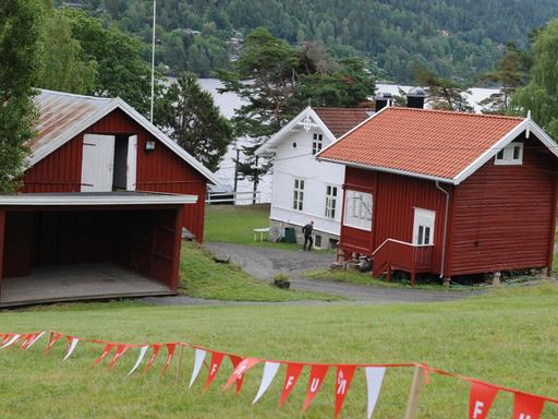 Drei Gebäude auf der norwegischen Insel Utøya. Im Vordergrund flattert ein rot-weißes Absperrband mit den Buchstaben AUF. Sie stehen für die Jugendorganisation der norwegischen Arbeiterpartei. Im Hintergrund sind Wasser und Bäume zu sehen.