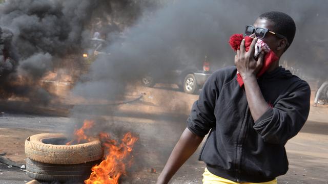 Ein Mann hält sich ein Tuch vor den Mund, im Hintergrund brennen Reifen.