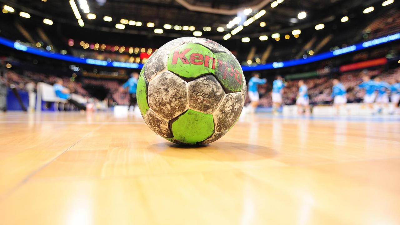 Das Bild zeigt einen schmutzigen grün-weißen Handball, der auf dem Parkett des Platzes liegt. Im Hintergrund unscharf Spieler und die Zuschauerränge. Das Bild zeigt einen schmutzigen grün-weißen Handball, der auf dem Parkett des Platzes liegt. Im Hintergrund unscharf Spieler und die Zuschauerränge.