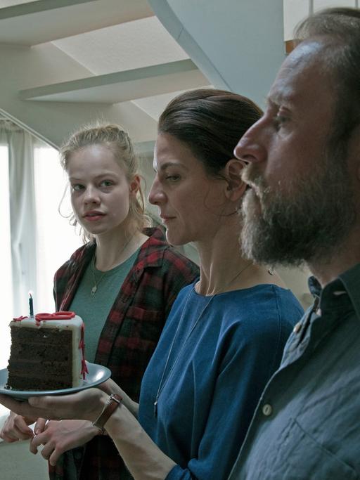 Szene aus "1000 Arten Regen zu beschreiben" von Isa Prahl: Schwester Miriam (Emma Bading, v. l. n. r.) und die Eltern Susanne (Bibiana Beglau) und Thomas (Bjarne Mädel) stehen mit dem Geburtstagskuchen vor der Tür zu Mikes Zimmer.