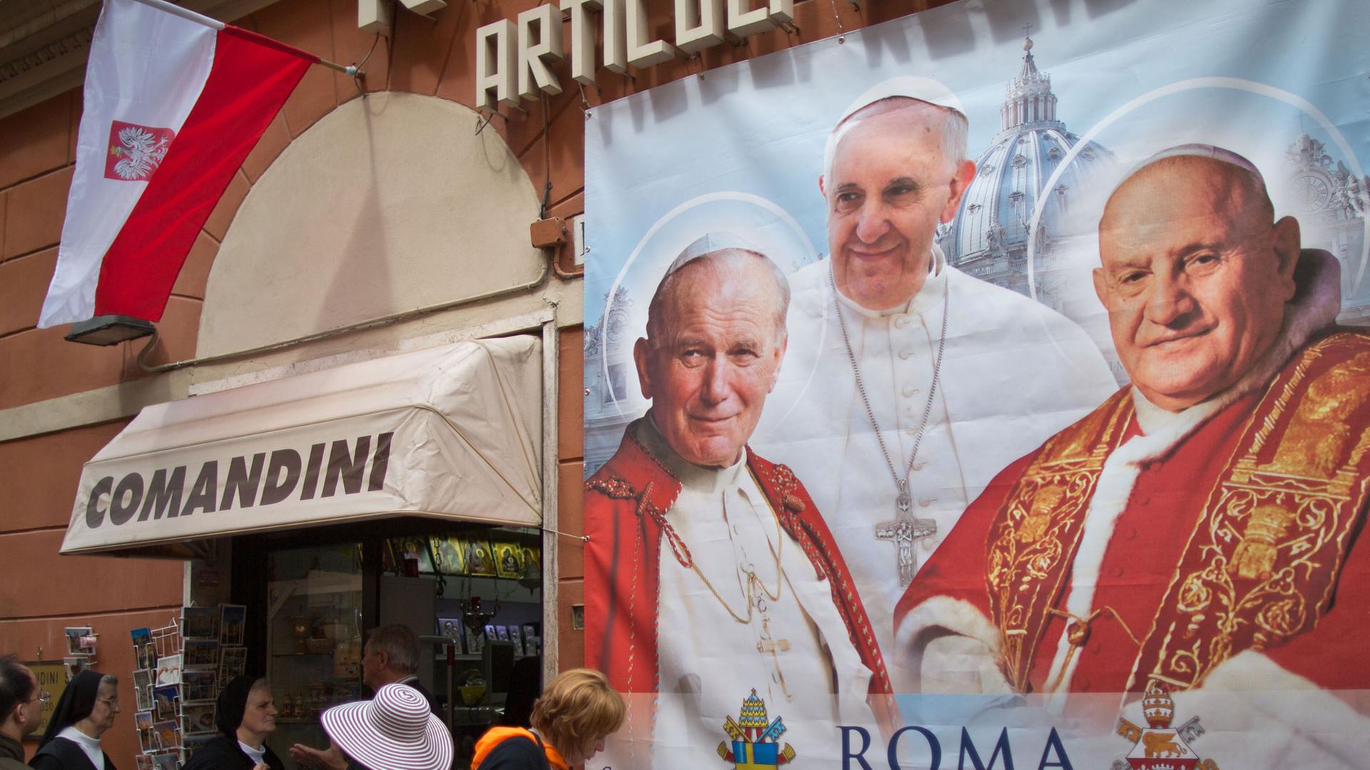 Menschen stehen vor einem Plakat mit den Päpsten Franziskus, Johannes XXIII. und Johannes Paul II.