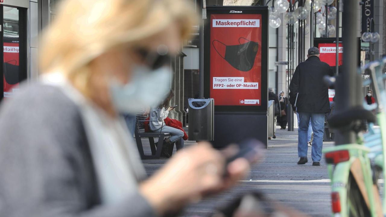 Eine Frau sitzt mit Maske in der Fußgängerzone vor einer Anzeigetafel, die auf die Maskenpflicht hinweist.