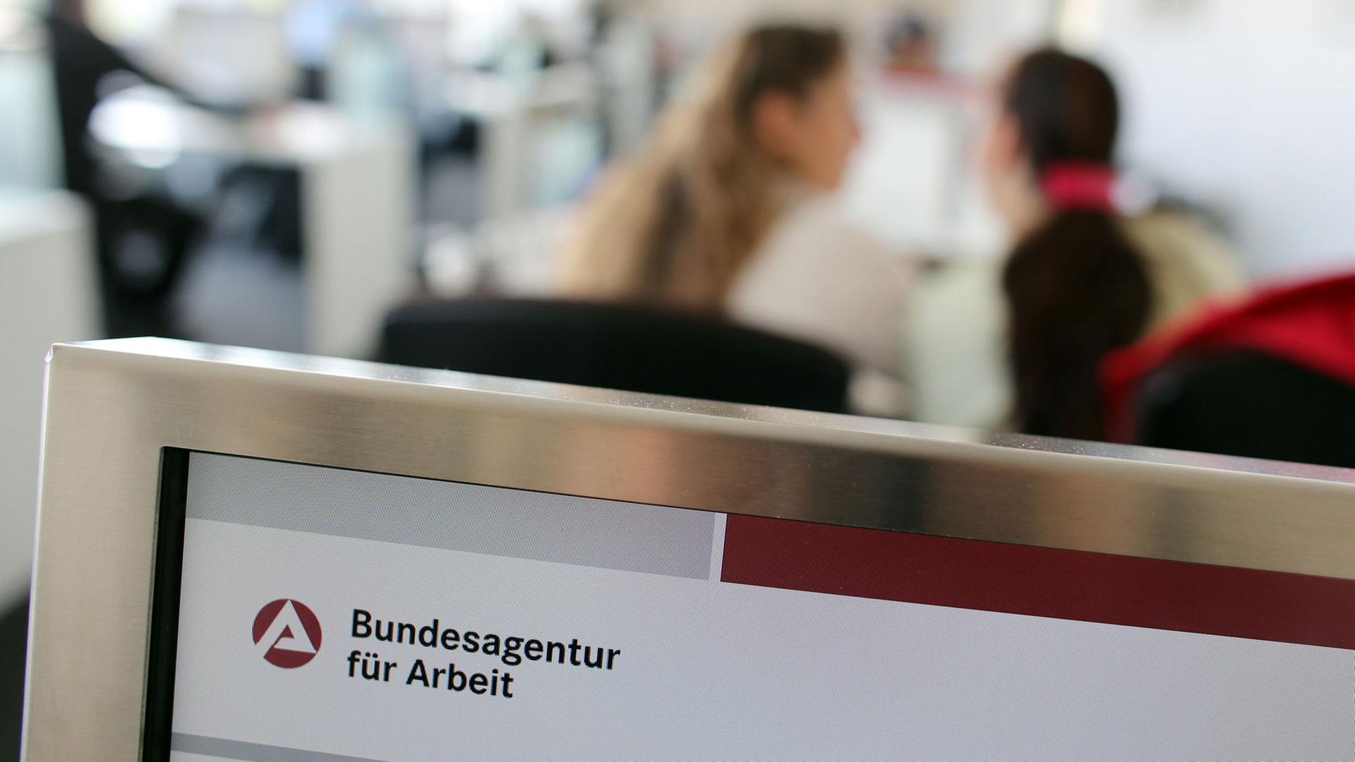 Blick in ein Jobcenter: im Vordergrund das Logo der Bundesagentur für Arbeit, im Hintergrund verschwommen zwei Frauen, die miteinander reden Blick in ein Jobcenter: im Vordergrund das Logo der Bundesagentur für Arbeit, im Hintergrund verschwommen zwei Frauen, die miteinander reden
