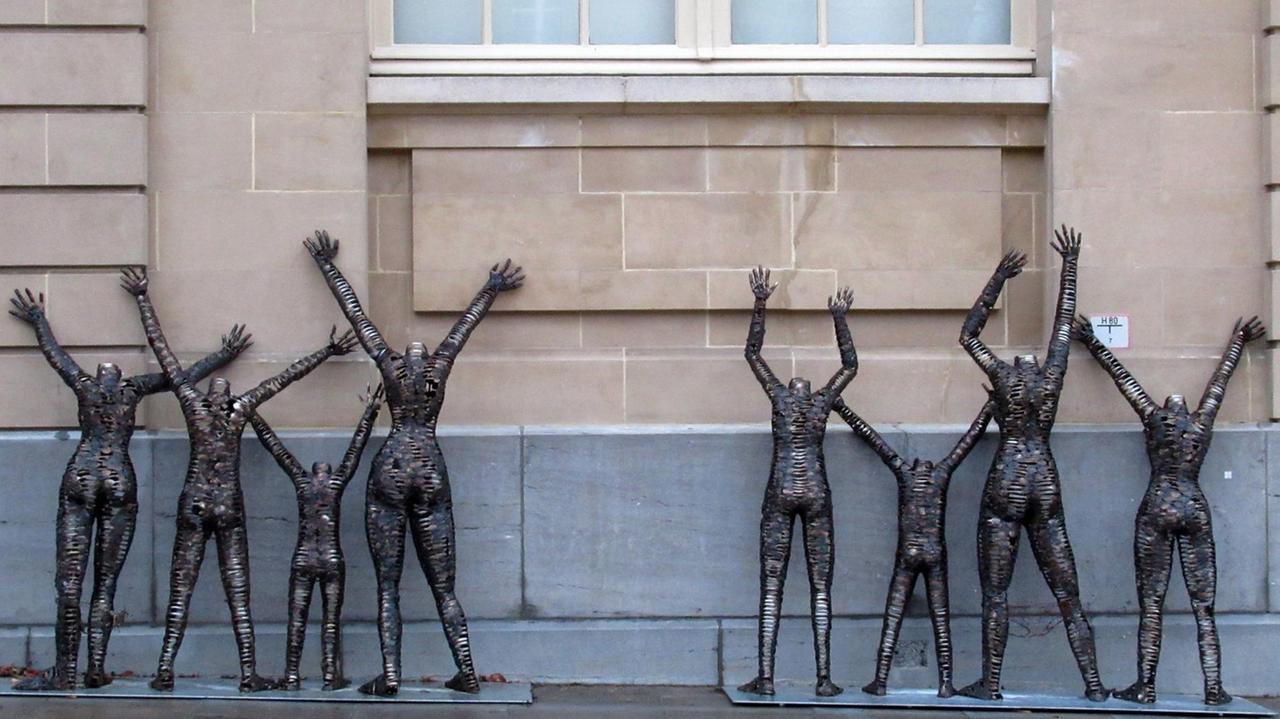 Graue Skulptur recken die Arme nach oben und stehen an einer Außenwand des Museums. Graue Skulptur recken die Arme nach oben und stehen an einer Außenwand des Museums.