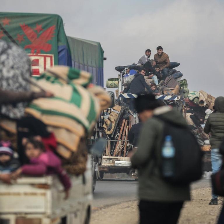 Syrer sitzen mit ihren Habseligkeiten auf vollbepackten Ladeflächen von Lastwagen. Im Vordergrund laufen zwei Männer auf ein Auto zu. Sie tragen Rucksäcke.