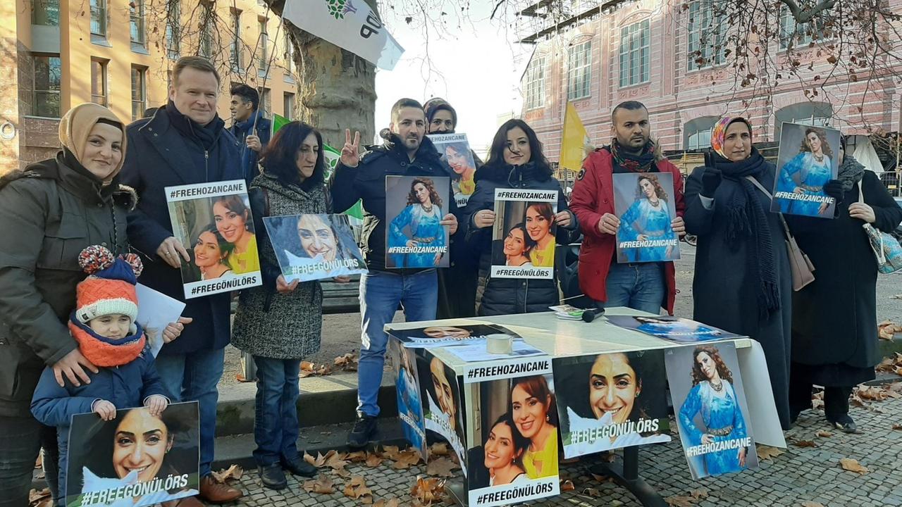Eine Gruppe von Menschen steht hinter einem Tisch unter einem Baum und hält Plakate hoch. Eine Gruppe von Menschen steht hinter einem Tisch unter einem Baum und hält Plakate hoch.