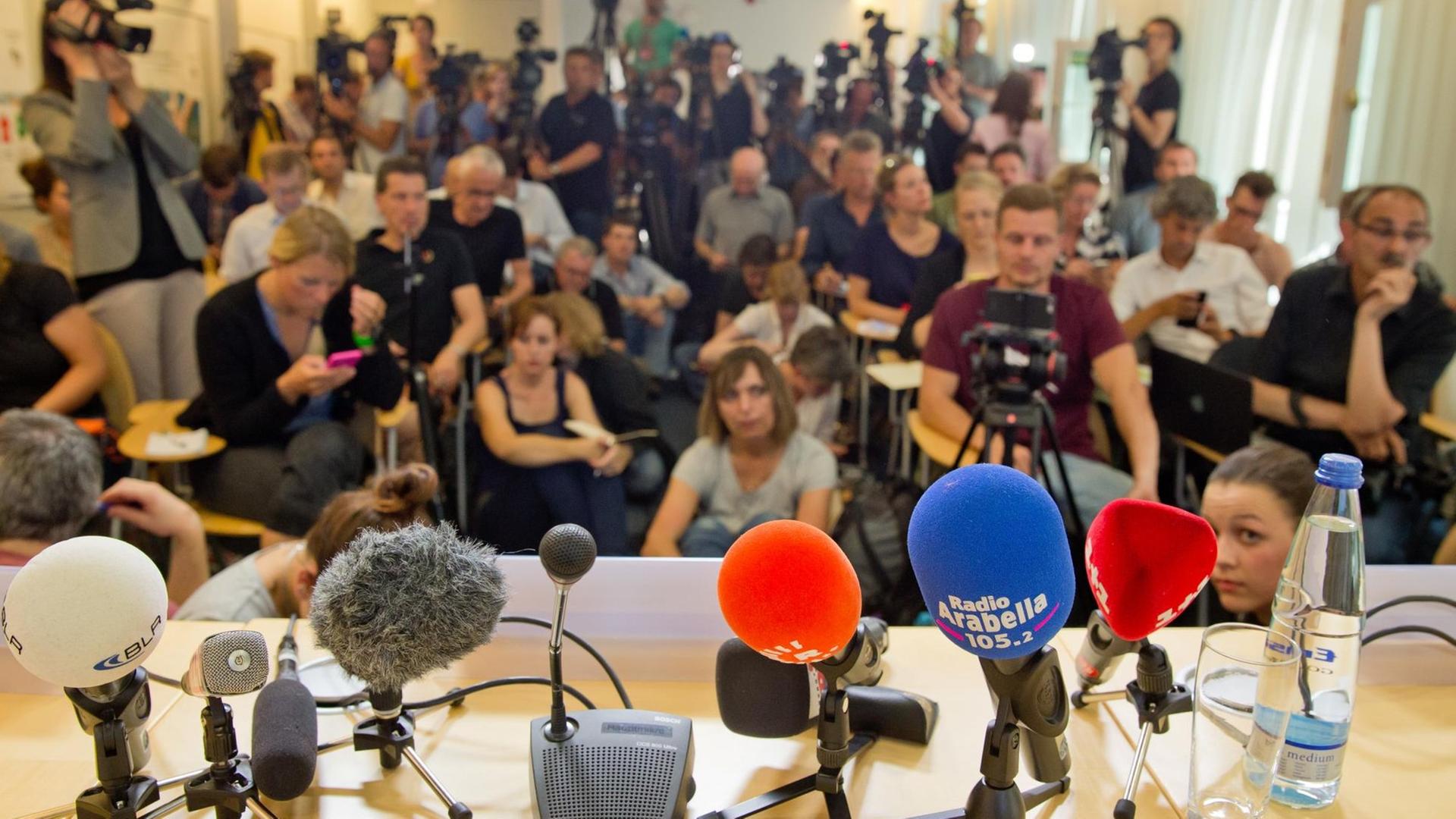 Sie sehen bunte Mikrofone und im Hintergrund viele Journalisten.