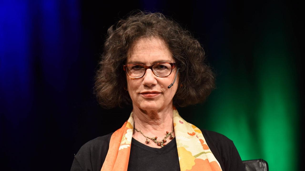 Susan Neiman, braune Locken, Brille, trägt ein schwarzes Oberteil und orange-gelben Schal mit Blumen. Sie sitzt vor einem schweren schwarzen Vorhang, der blau und grün beleuchtet ist. Susan Neiman, braune Locken, Brille, trägt ein schwarzes Oberteil und orange-gelben Schal mit Blumen. Sie sitzt vor einem schweren schwarzen Vorhang, der blau und grün beleuchtet ist.
