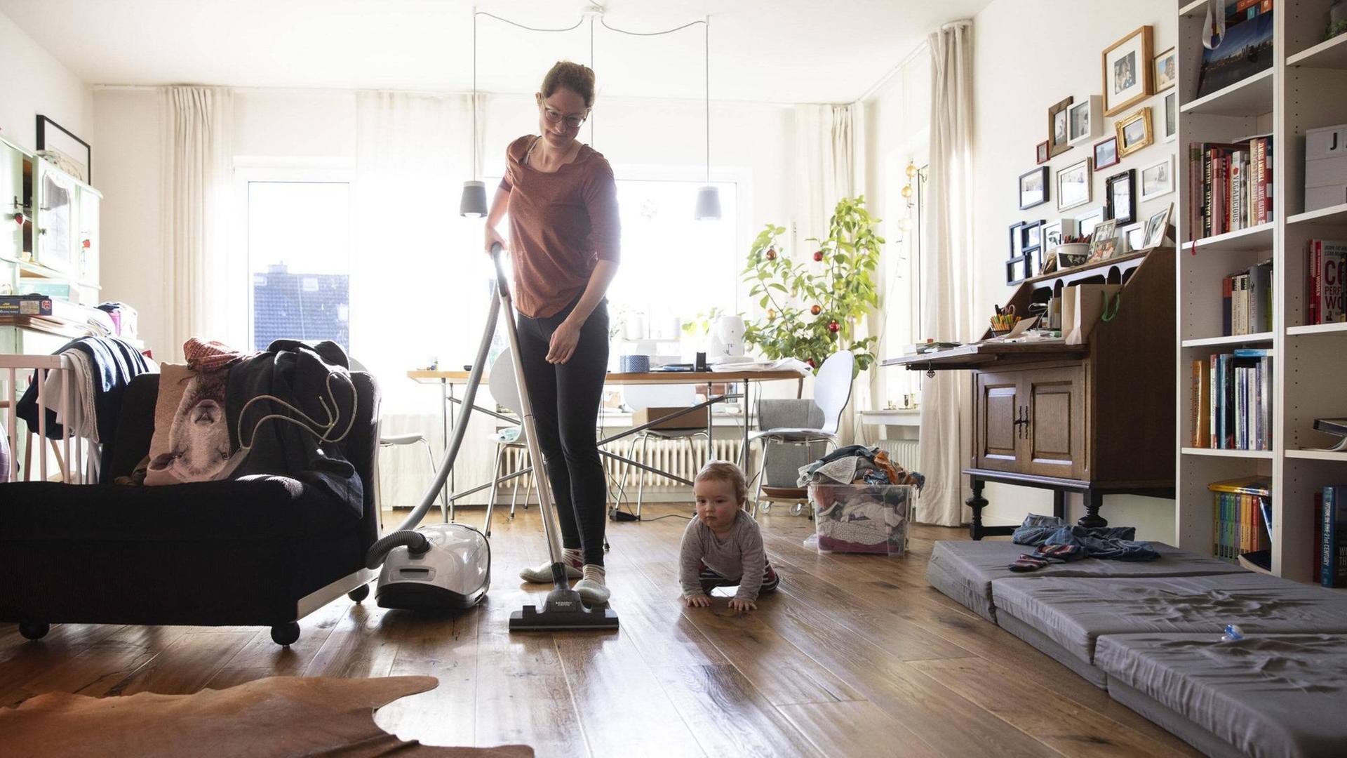 Eine Frau saugt in ihrem Wohnzimmer Staub, am Boden sitzt ein Kleinkind. Eine Frau saugt in ihrem Wohnzimmer Staub, am Boden sitzt ein Kleinkind.
