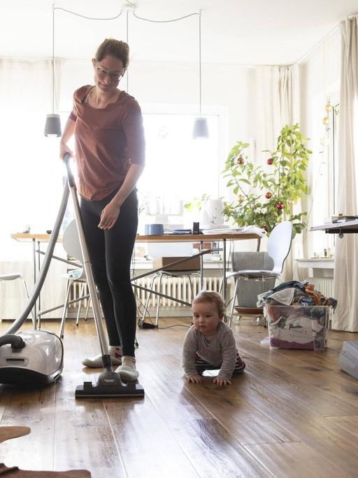 Eine Frau saugt in ihrem Wohnzimmer Staub, am Boden sitzt ein Kleinkind.