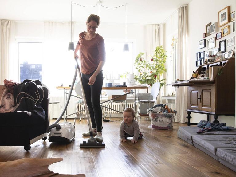 Eine Frau saugt in ihrem Wohnzimmer Staub, am Boden sitzt ein Kleinkind.