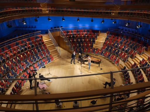 Saalansicht eines geschwungenen Konzertraumes mit roten Sesseln, die ovalförmig um die Bühne angeordnet sind. Auf dem Parkett steht ein Flügel, mehrere Menschen und zwei Kameras.