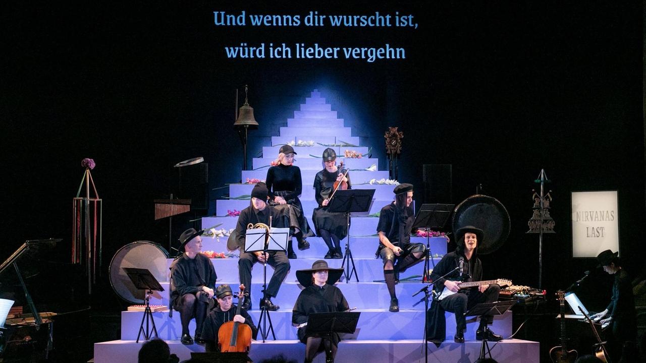 Das Ensemble der Münchner Kammerspiele bei der Aufführung "Nirvanas Last" auf der Bühne auf einer blauen Treppe sitzend. Die Schauspieler stellen Musiker in einer Kammerformation dar. Über ihnen ist ein Schriftzug zu lesen: "Und wenns dir wurscht ist, würd ich lieber vergehn".
