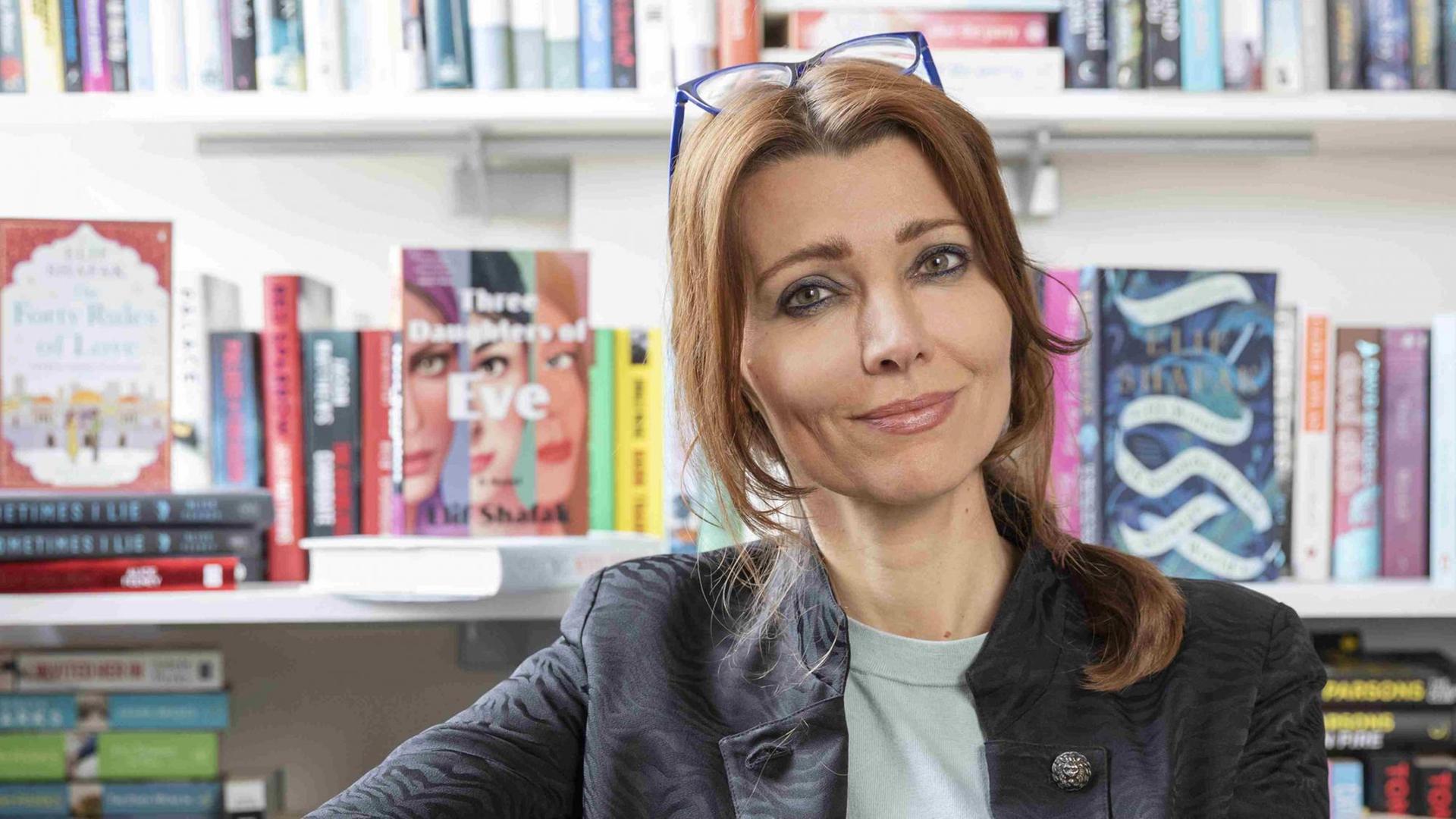 Elif Shafak sitzt vor einem Bücherregal und schaut freundlich in die Kamera. Elif Shafak sitzt vor einem Bücherregal und schaut freundlich in die Kamera.