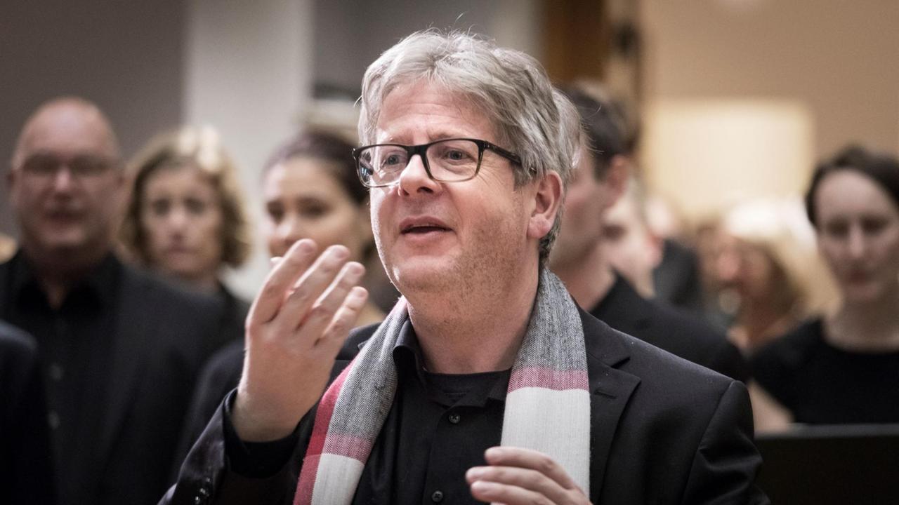 Ein grauhaariger Mann mit Brille schaut konzentriert nach links und gestikuliert mit den Händen - im Hintergrund weitere Menschen in schwarzer Kleidung, Ein grauhaariger Mann mit Brille schaut konzentriert nach links und gestikuliert mit den Händen - im Hintergrund weitere Menschen in schwarzer Kleidung,