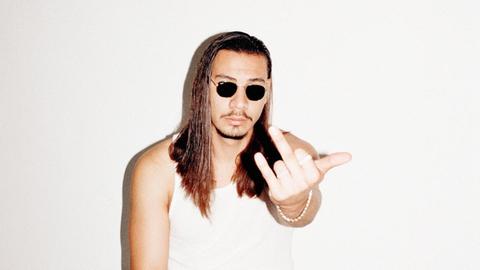 Ein Promotionfoto zeigt den Deutschrapper Apache207 direkt angeblitzt mit Sonnenbrille vor weißer Wand und scenetypischer Geste.