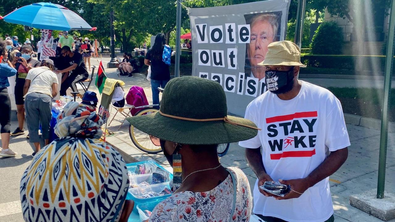 Menschen bei einer Kundgebung von Black Lives Matter in Washington D.C. An einem Stand verteilt ein Mann mit "Stay Woke"-T-Shirt Flyer. Menschen bei einer Kundgebung von Black Lives Matter in Washington D.C. An einem Stand verteilt ein Mann mit "Stay Woke"-T-Shirt Flyer.