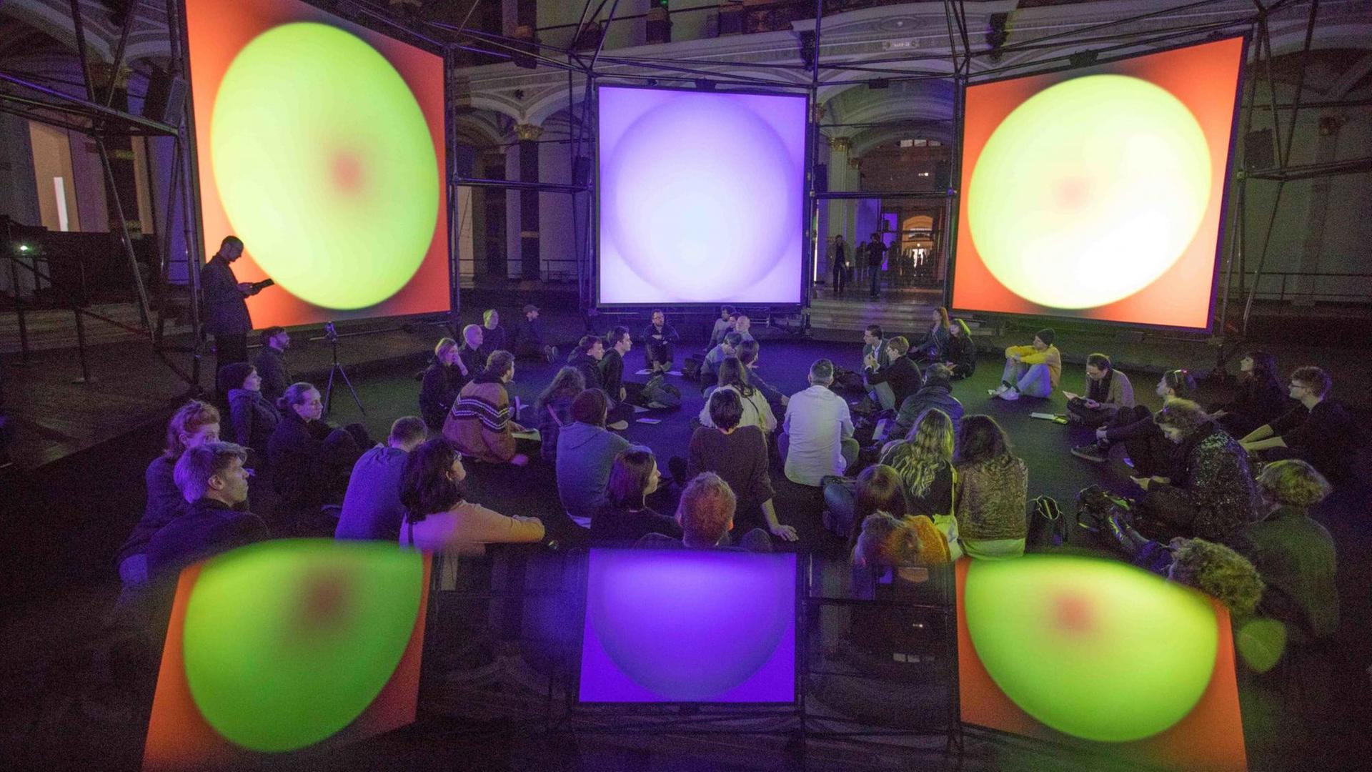 Spiegelung der Installation "Empty Formalism" von Brian Eno im "ISM Hexadome" im Martin-Gropius-Bau. Die Installation zeigt auf Leinwänden farbige, sich kontinuierlich wandelnde Flächen, dazu sind sphärische Klänge zu hören.