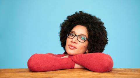 Kenza Ait Si Abbou blickt selbstbewußt mit Brille und rotem Pullover auf einem Holztisch aufgestützt und vor einem blauen Hintergrund den Betrachter an.