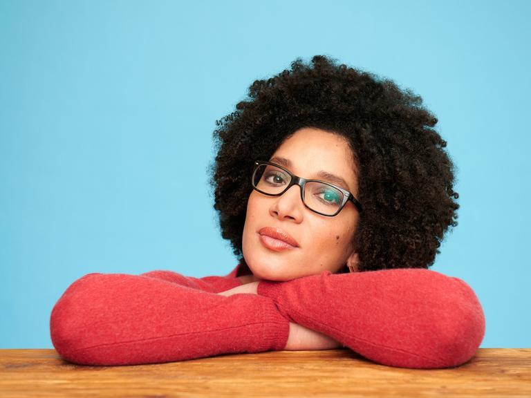 Kenza Ait Si Abbou blickt selbstbewußt mit Brille und rotem Pullover auf einem Holztisch aufgestützt und vor einem blauen Hintergrund den Betrachter an.