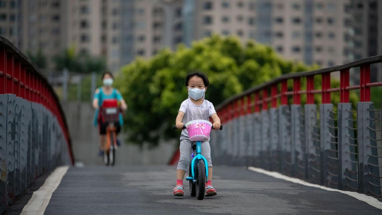 Ein taiwanisches Kind mit einer Schutzmaske fährt mit einem Fahrrad über eine Brücke. Im Hintergrund ist eine erwachsene Person zu sehen.
