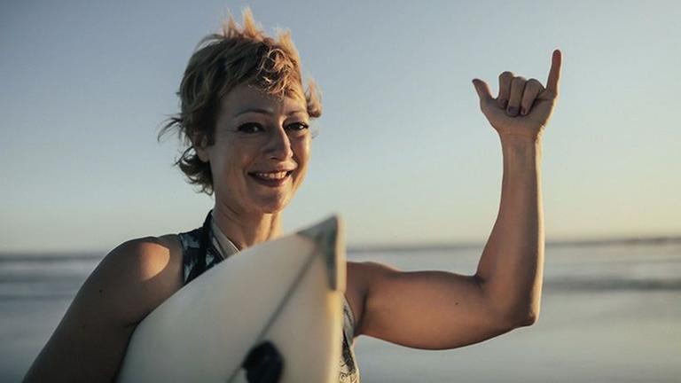 Frau mit Surfboard, die eine Geste mit Daumen und kleinem Finger Richtung Kamera macht.