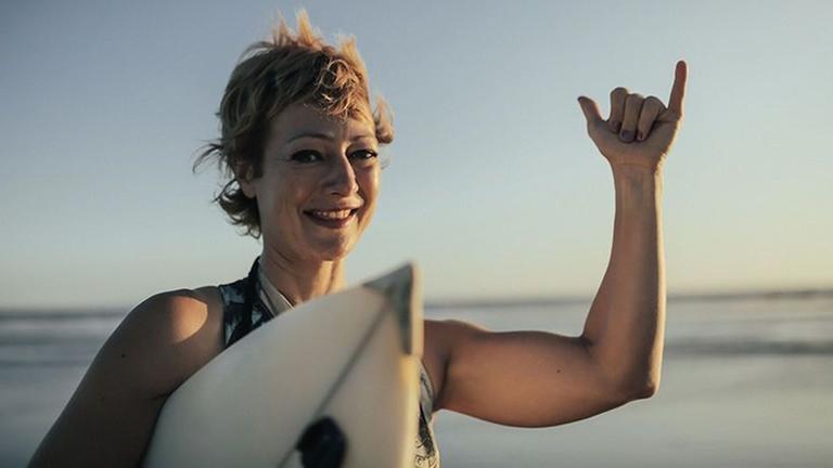 Frau mit Surfboard, die eine Geste mit Daumen und kleinem Finger Richtung Kamera macht. Frau mit Surfboard, die eine Geste mit Daumen und kleinem Finger Richtung Kamera macht.