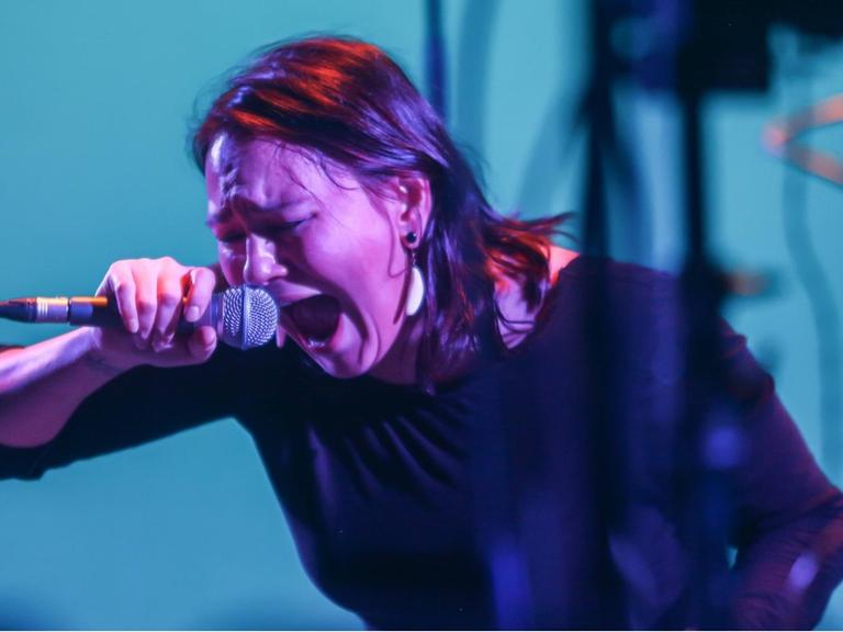 Tanya Tagaq steht auf einer blau erleuchteten Bühne in violettem Licht und schreit in das Mikrofon.