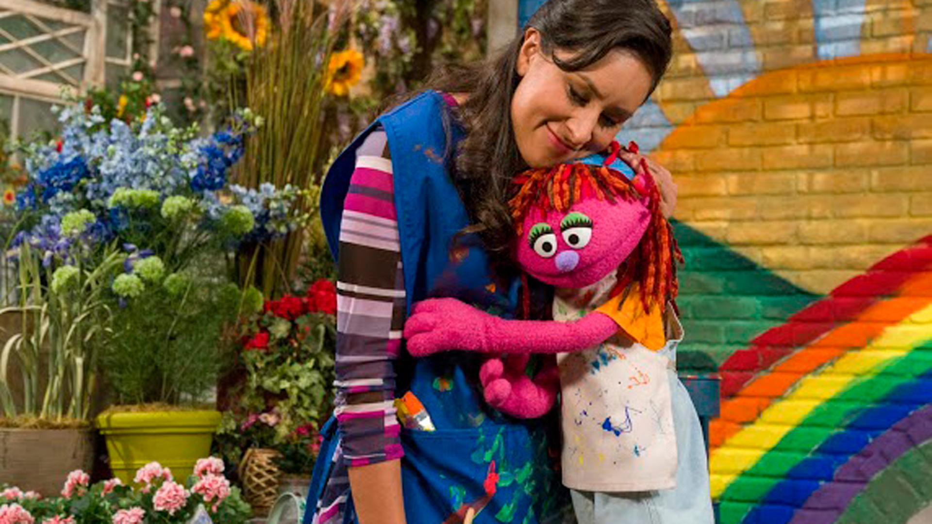 Die Puppe Lily aus der Fernsehserie Sesamstraße, die mit ihrer Familie obdachlos wird, schmiegt sich an eine Frau.