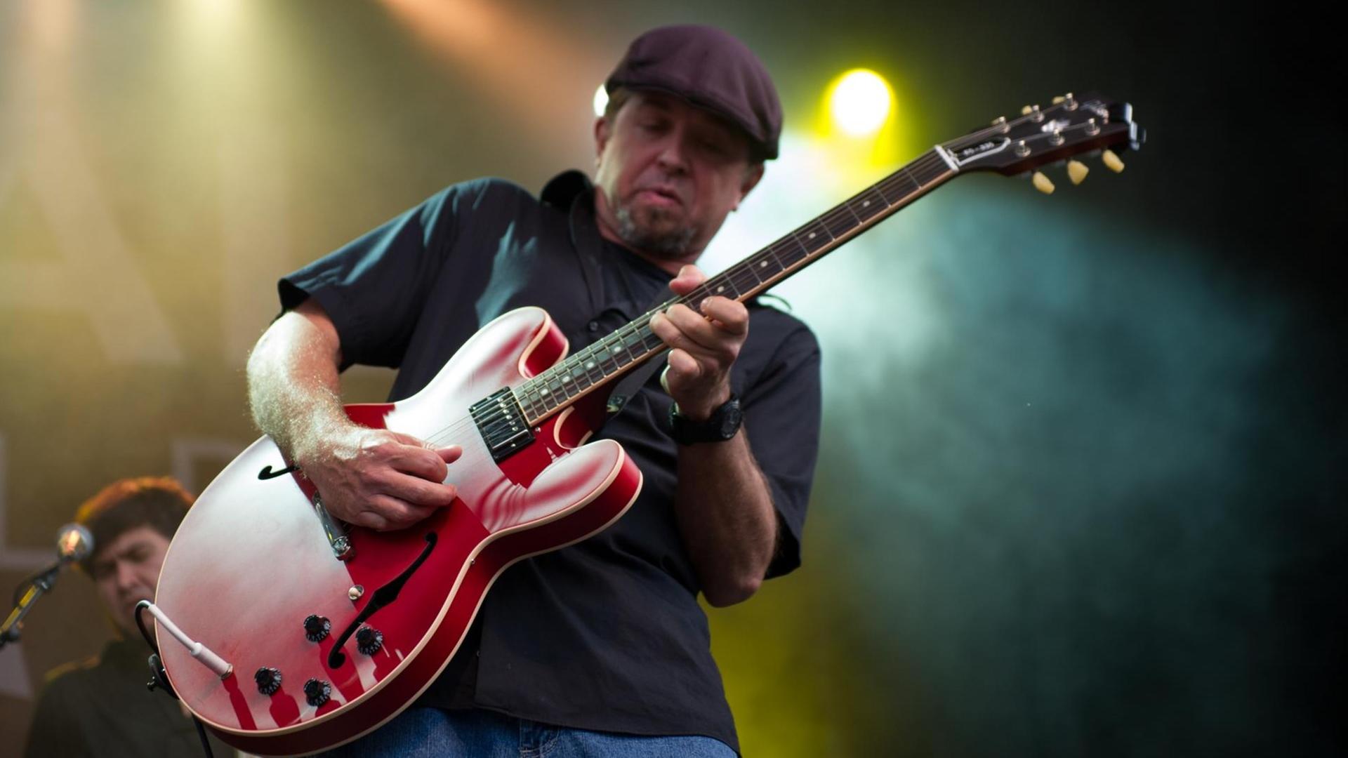 Ein Mann mit Kappe steht auf einer Bühne und spielt eine rote Gitarre. Ein Mann mit Kappe steht auf einer Bühne und spielt eine rote Gitarre.