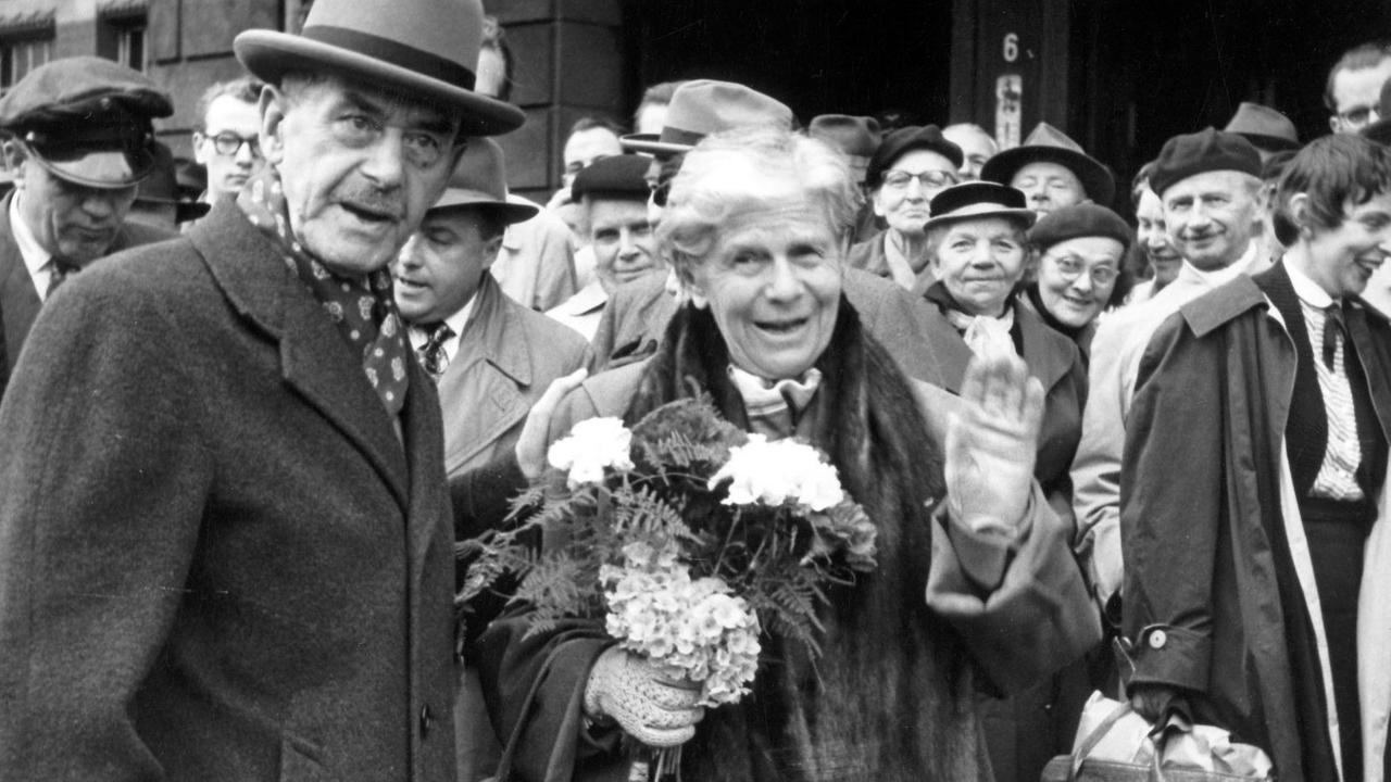 Der deutsche Schriftsteller Thomas Mann mit seiner Ehefrau Katja, die in die Kamera winkt. Hinter ihnen stehen viele Menschen. Der deutsche Schriftsteller Thomas Mann mit seiner Ehefrau Katja, die in die Kamera winkt. Hinter ihnen stehen viele Menschen.