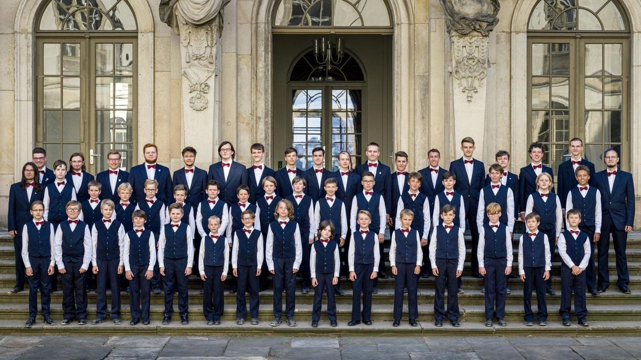 Der Knabenchor steht in blauen Westen auf den Stufen vor einem schlossähnlichen Gebäude. Der Knabenchor steht in blauen Westen auf den Stufen vor einem schlossähnlichen Gebäude.
