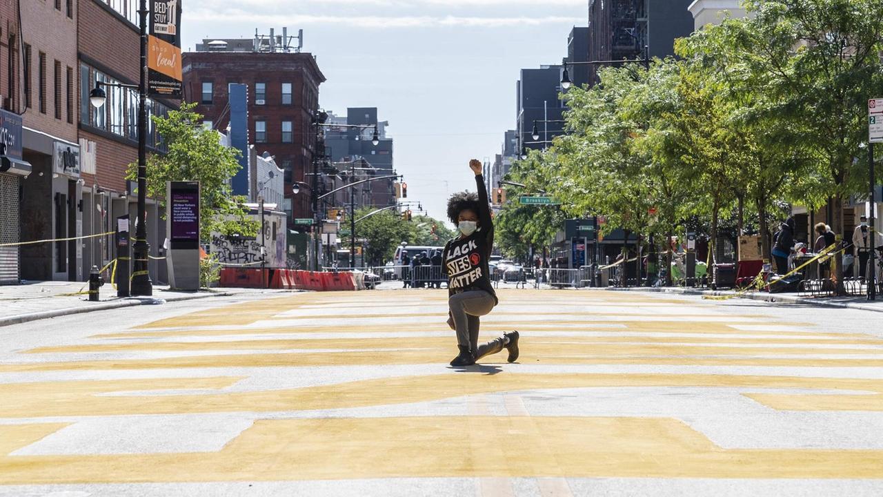 Eine Black Lives Matter-Aktivistin während einer Kunst- und Protestaktion in New York. Sie kniet auf einem Knie auf einer leeren Straße und reckt eine Faust zum Himmel.