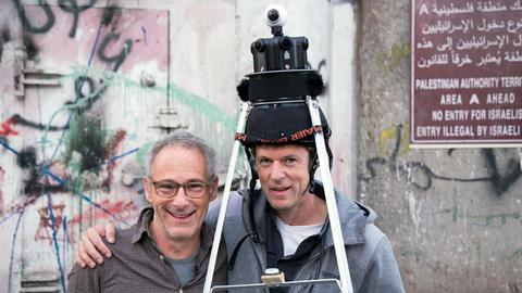 Regisseur Dani Levy und Kameramann Filip Zumbrunn am Drehort der Episode "Liebe/Love" vor der Sperrmauer. Zumbrunn hat ein auffälliges Gerüst auf dem Kopf, in dem sich seine Kamera befindet. Beide lachen in die Kamera. Zumbrunn umarmt Levy.