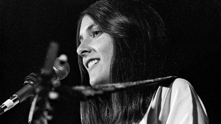 Schwarz-Weiß-Bild von Mimi Fariña auf der Bühne mit einer Gitarre in der Hand. Schwarz-Weiß-Bild von Mimi Fariña auf der Bühne mit einer Gitarre in der Hand.