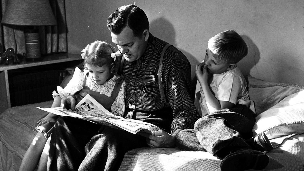 Ein Vater liest auf einem Bett sitzend seinen Kindern vor.